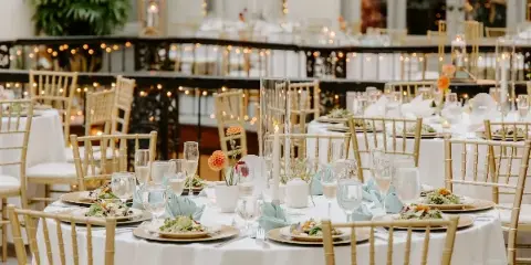 A wedding reception venue featuring round tables with white linens, gold chiavari chairs, and floral centerpieces.