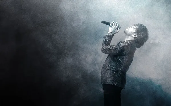 A male singer performing on stage with a microphone in a smoky, atmospheric setting.