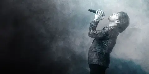 A male singer performing on stage with a microphone in a smoky, atmospheric setting.
