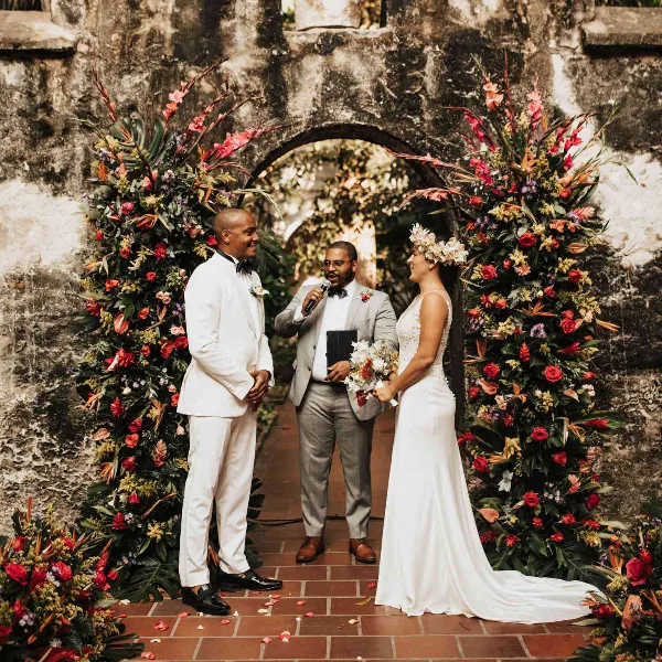 A wedding officiant in a grey suit holding a microphone and book, standing between a bride and groom at a floral arch.