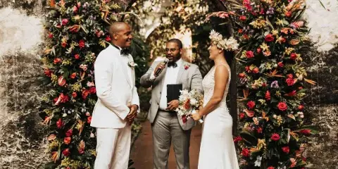 A wedding officiant in a grey suit holding a microphone and book, standing between a bride and groom at a floral arch.
