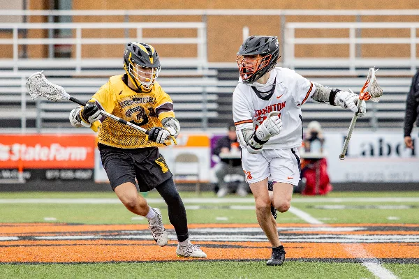 Two lacrosse players in action on a field, one in a yellow jersey and one in a white jersey, with stadium seating in the background.