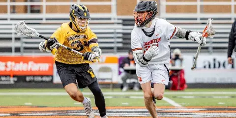 Two lacrosse players in action on a field, one in a yellow jersey and one in a white jersey, with stadium seating in the background.