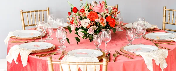Styled event table with coral linen, gold chiavari chairs, white plates, gold flatware, glassware, and a floral centerpiece.