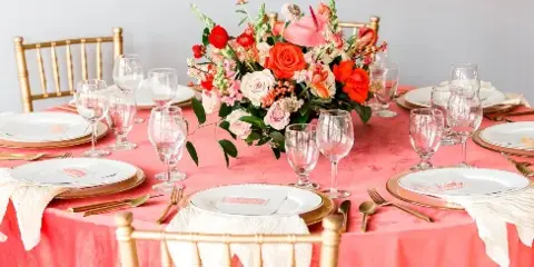 Styled event table with coral linen, gold chiavari chairs, white plates, gold flatware, glassware, and a floral centerpiece.
