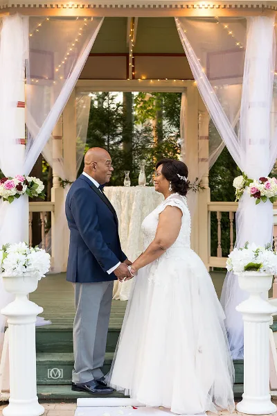 A bride and groom standing at a wedding altar on a decorated porch with white drapery and floral arrangements.