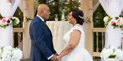 A bride and groom standing at a wedding altar on a decorated porch with white drapery and floral arrangements.