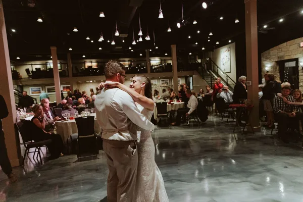A bride and groom dancing at a wedding reception in a banquet hall with seated guests and hanging lights.