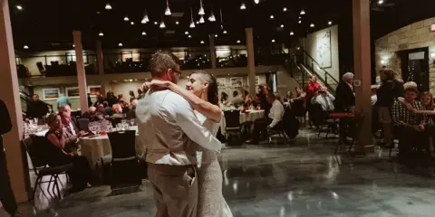 A bride and groom dancing at a wedding reception in a banquet hall with seated guests and hanging lights.