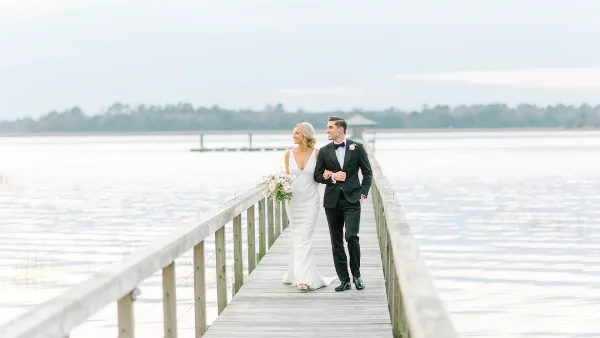 A bride and groom walking arm-in-arm on a wooden pier over a body of water.