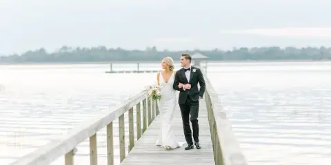 A bride and groom walking arm-in-arm on a wooden pier over a body of water.