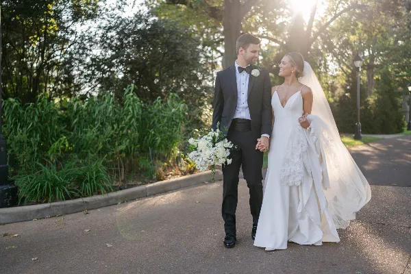 A bride and groom walking hand-in-hand on a paved path outdoors, with the bride holding a white bouquet and wearing a long veil.