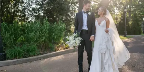 A bride and groom walking hand-in-hand on a paved path outdoors, with the bride holding a white bouquet and wearing a long veil.