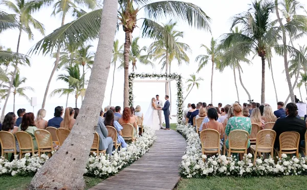 A bride and groom exchanging vows at a beachside wedding ceremony under a floral arch, with seated guests and palm trees in the background.
