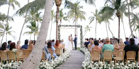 A bride and groom exchanging vows at a beachside wedding ceremony under a floral arch, with seated guests and palm trees in the background.