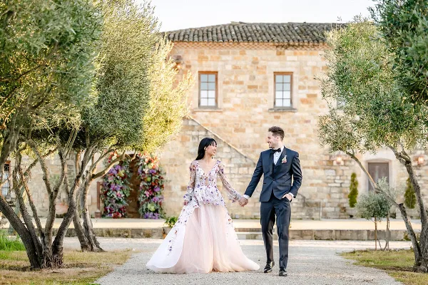 A bride and groom holding hands and walking outdoors in front of a stone building with olive trees.