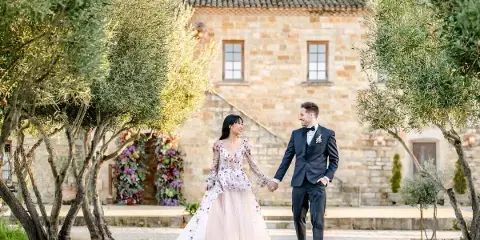 A bride and groom holding hands and walking outdoors in front of a stone building with olive trees.