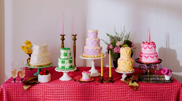 A table display featuring several vintage-style tiered wedding cakes in various colors and piping styles, accompanied by floral arrangements and candles.