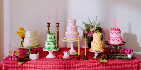 A table display featuring several vintage-style tiered wedding cakes in various colors and piping styles, accompanied by floral arrangements and candles.