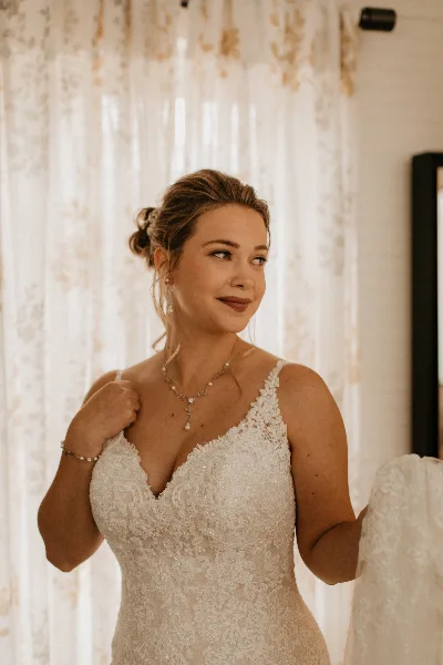A bride in a white lace wedding dress with an elegant updo hairstyle and professional makeup, standing in a bridal suite.