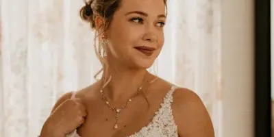 A bride in a white lace wedding dress with an elegant updo hairstyle and professional makeup, standing in a bridal suite.