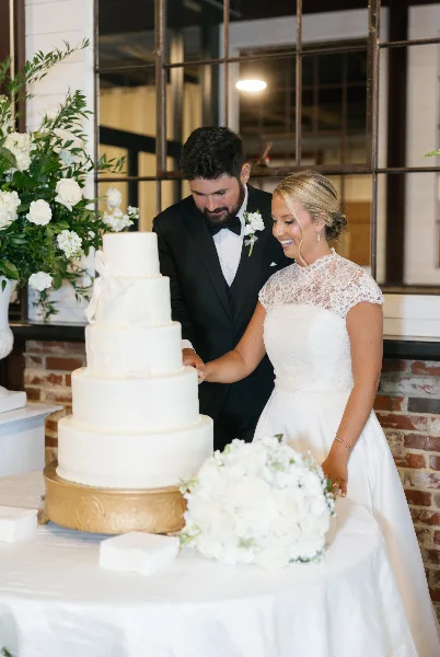 A bride and groom in formal attire cutting a white tiered wedding cake at a reception venue.