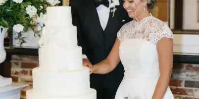 A bride and groom in formal attire cutting a white tiered wedding cake at a reception venue.