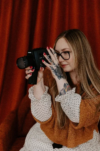 A woman with glasses and tattoos holding a vintage film camera against a red curtain background.