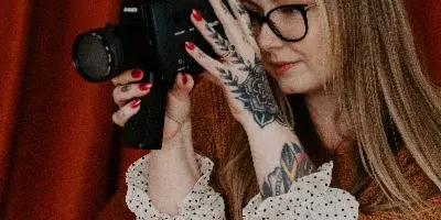 A woman with glasses and tattoos holding a vintage film camera against a red curtain background.