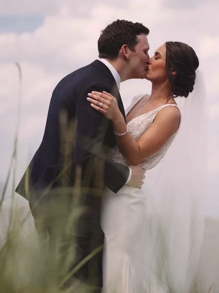 A bride and groom sharing a kiss in an outdoor setting, with the bride wearing a white gown and veil and the groom in a dark suit.