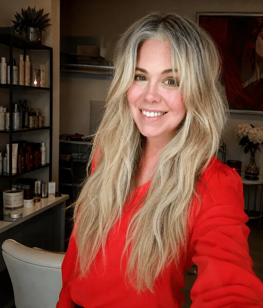 Portrait of a smiling woman with long blonde hair wearing a red top, standing in a beauty salon with shelves of hair products in the background.