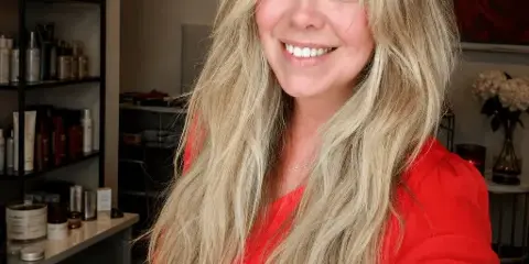 Portrait of a smiling woman with long blonde hair wearing a red top, standing in a beauty salon with shelves of hair products in the background.