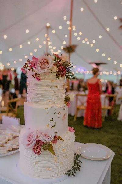 A three-tiered white wedding cake with textured frosting, fresh pink roses, and gold leaf accents, displayed on a table at a wedding reception.