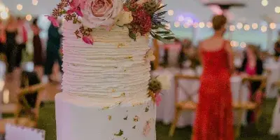 A three-tiered white wedding cake with textured frosting, fresh pink roses, and gold leaf accents, displayed on a table at a wedding reception.