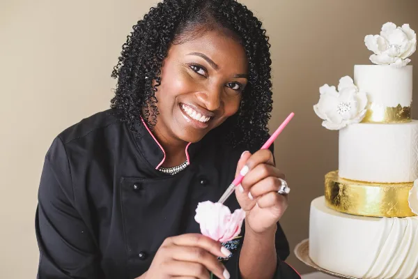 A smiling baker in a black chef's coat holding a piping tool, standing next to a decorated three-tier white wedding cake with gold bands and sugar flowers.