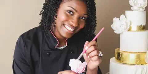A smiling baker in a black chef's coat holding a piping tool, standing next to a decorated three-tier white wedding cake with gold bands and sugar flowers.