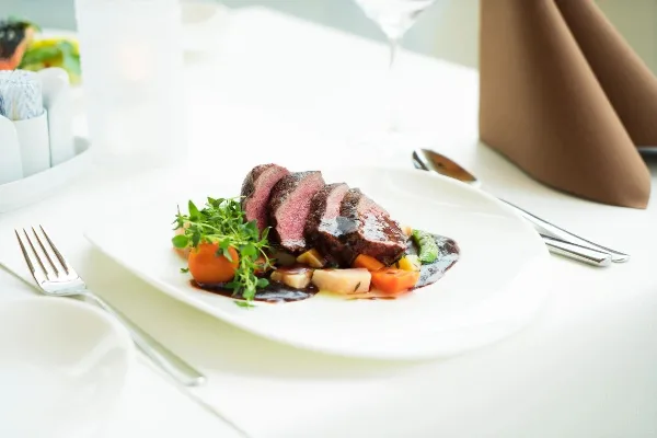 A plated sliced beef entrée with vegetables and sauce on a white table setting with silverware and folded napkins.