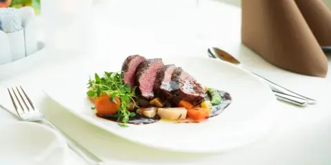 A plated sliced beef entrée with vegetables and sauce on a white table setting with silverware and folded napkins.