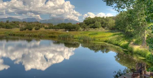A scenic view of a calm lake reflecting the sky and mountains, surrounded by green meadows and trees at the Blue Lake Ranch venue.
