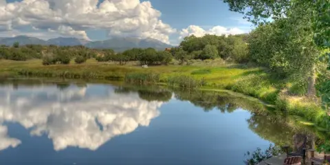 A scenic view of a calm lake reflecting the sky and mountains, surrounded by green meadows and trees at the Blue Lake Ranch venue.
