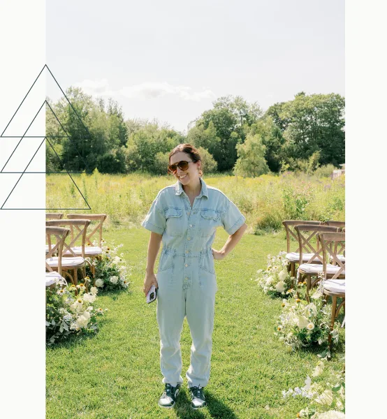 A woman in denim overalls standing in a grassy wedding aisle with chairs and flowers.