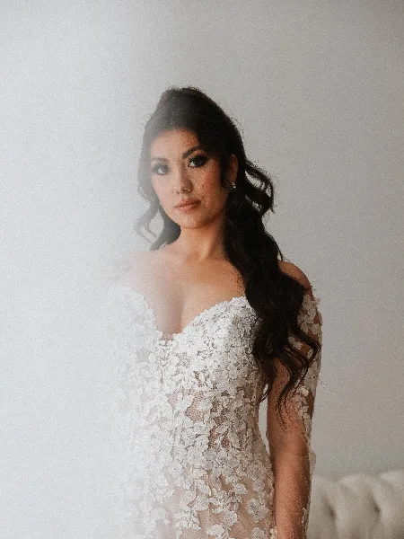 A bride wearing a white lace wedding dress with professionally styled wavy hair and full makeup.