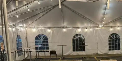 Interior of a white event tent set up with tables, chairs, and string lights.