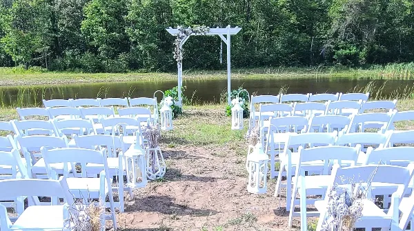 An outdoor wedding ceremony setup with white chairs and an arch facing a pond and wooded area.