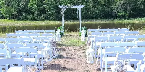An outdoor wedding ceremony setup with white chairs and an arch facing a pond and wooded area.