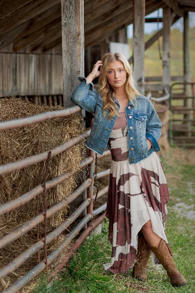 A young woman in a denim jacket and patterned dress posing beside a wooden barn fence with hay bales.
