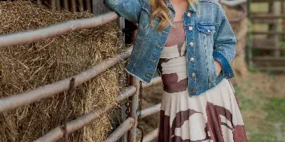 A young woman in a denim jacket and patterned dress posing beside a wooden barn fence with hay bales.