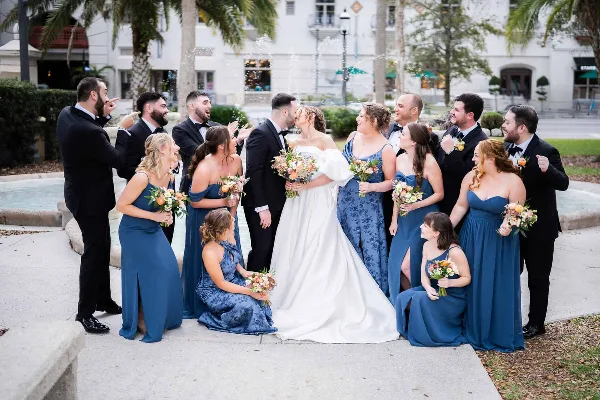 A wedding party, including the bride and groom, poses together outdoors near a fountain, with the couple kissing and the wedding party laughing and celebrating.