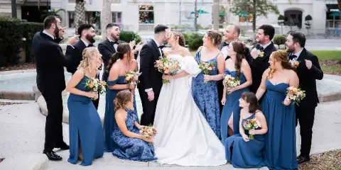 A wedding party, including the bride and groom, poses together outdoors near a fountain, with the couple kissing and the wedding party laughing and celebrating.