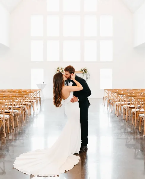A bride and groom kissing in a bright, white wedding reception hall with rows of wooden chairs.